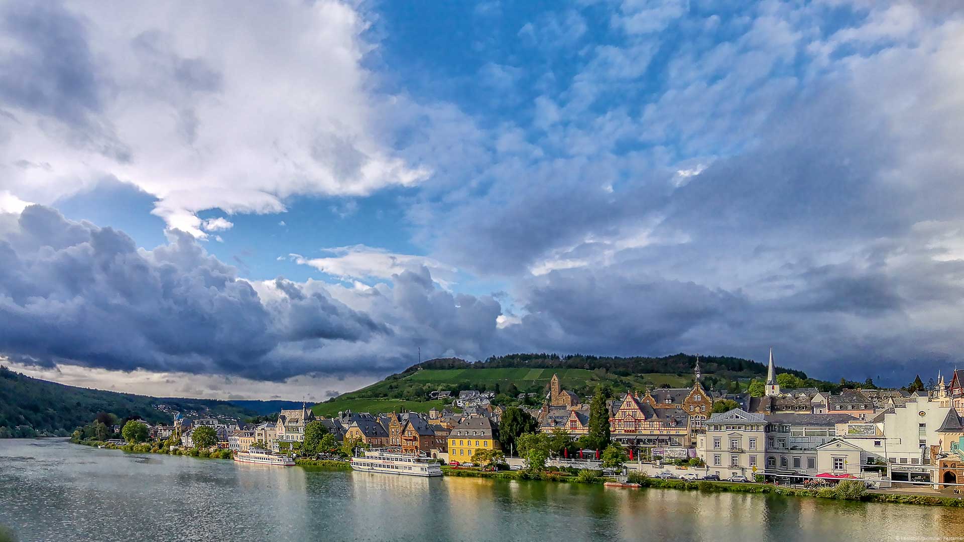 Vorne fließt der Fluss Mosel. Am gegenüberliegenden Ufer erkennt man viele Bauten im Stile des Jugendstils im Ort Traben-Trarbach. Am blauen Himmel sind mächtige Wolken. Im Hintergrund sind bewaldete Hügel und Weinberge.