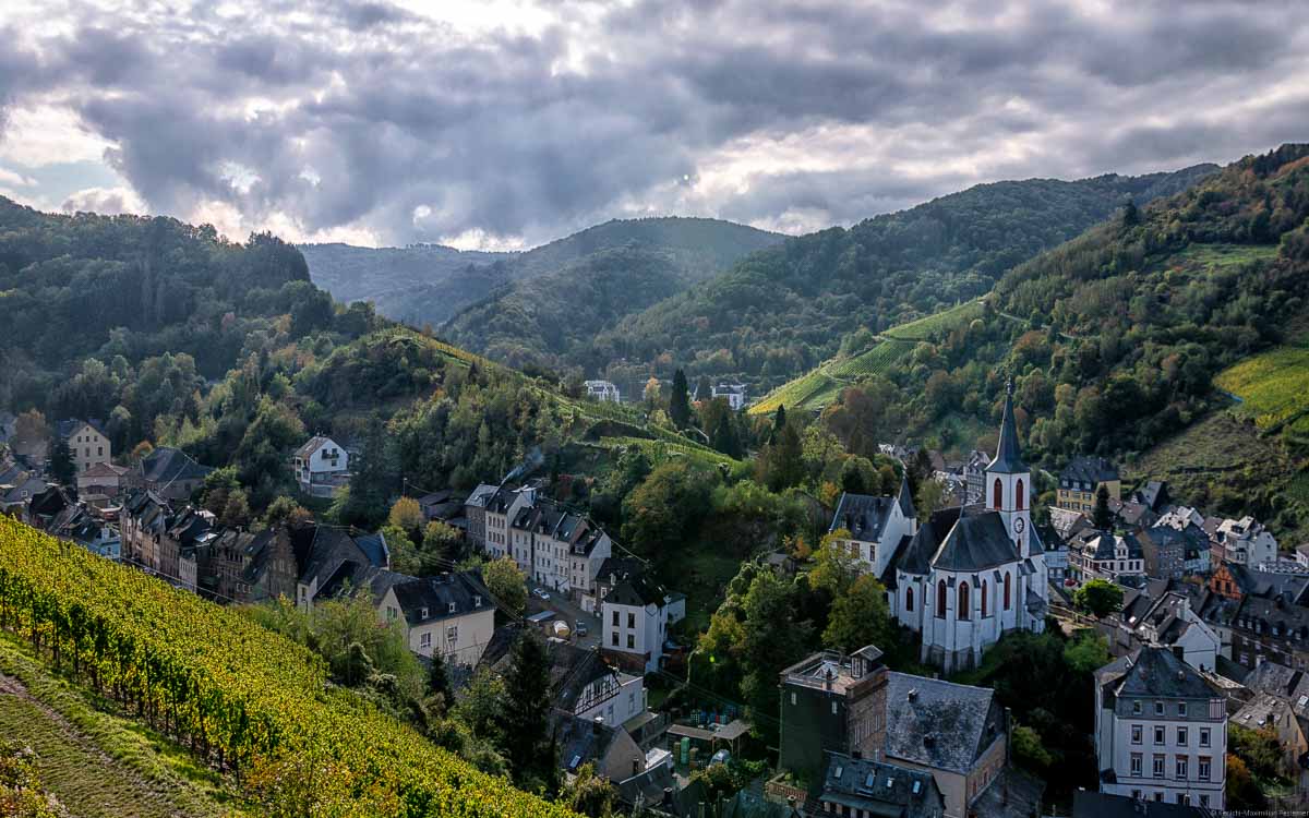 IVorne links befindet sich der Weinberg Trarbacher Burgberg. In der Mitte sieht man die Häuser und die Kapelle sowie die Pilgerherberge in Traben-Trarbach. Im Hintergrund rechts erkennt man den Weinberg Trarbacher Schlossberg. Von dort stammt der Traben-Trarbacher Wein. Im Hintergrund sind auch bewaldete Hügel. Am Himmel sind viele und große Wolken.