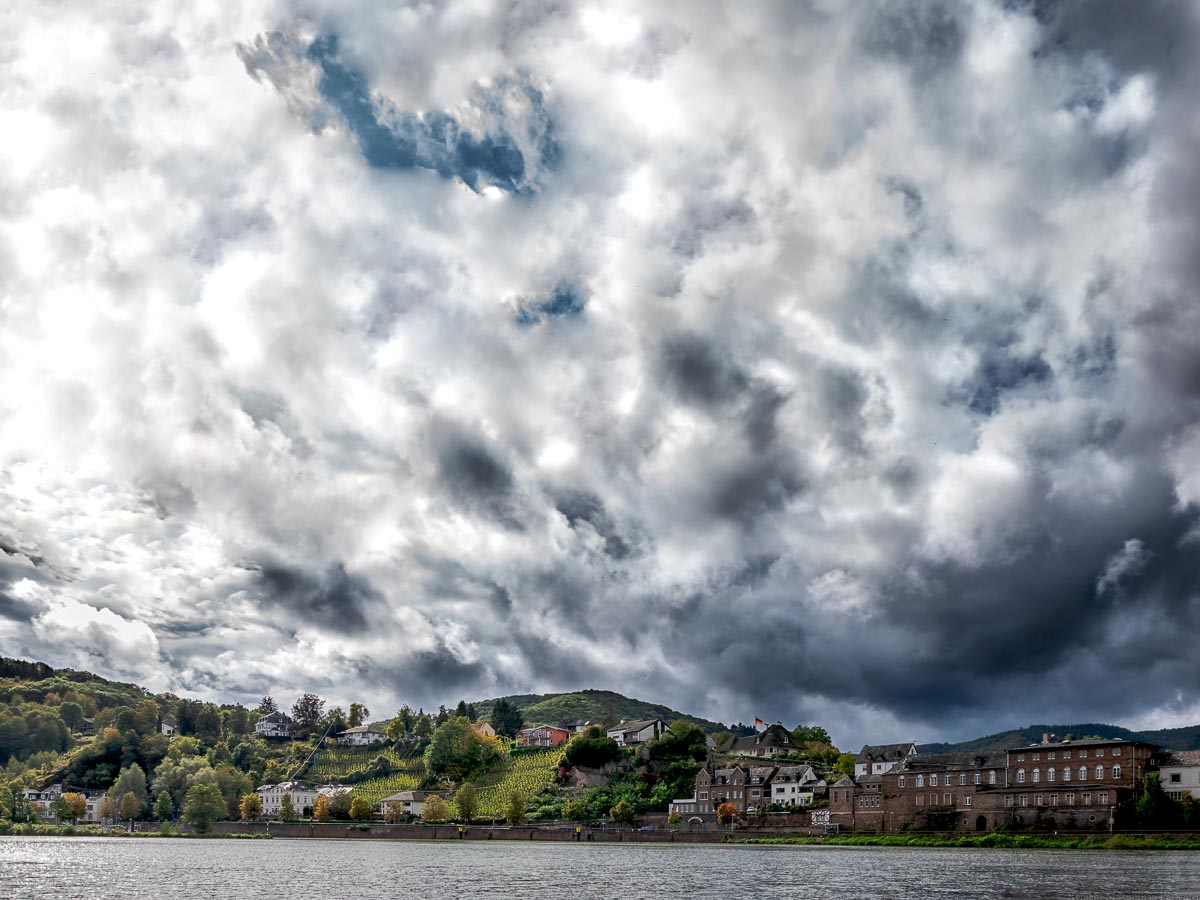 Im Vordergrund fließt der Fluss Mosel. Die Alfer Hoelle ist ein Weinberg der sich in den Ort Alf einfügt. Der Weinberg liegt an einem Hang zwischen Häusern am Ufer des Flusses. Der Himmel ist stark bewölkt.