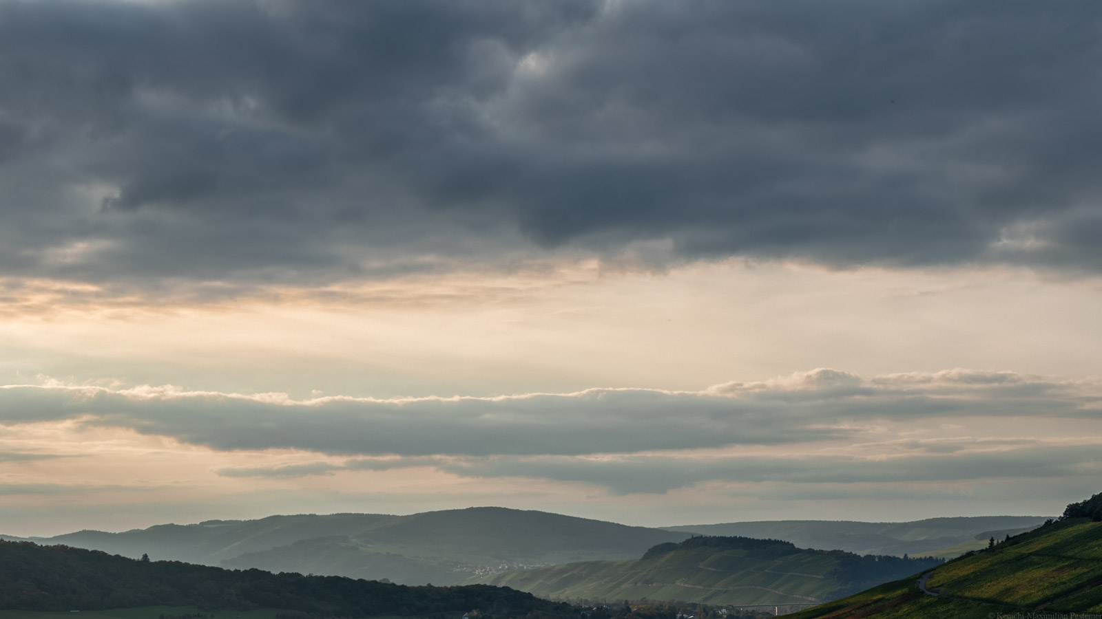 In der Mitte erkennt man den Weinberg Brauneberger Juffer Sonnenuhr am späten Abend. Links und rechts sind weitere Weinberge und bewaldete Hügel. Am Himmel sind blau-graue Wolken und eine orangene Färbung erkennbar.
