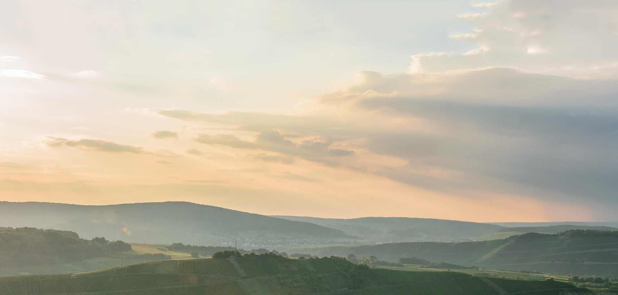 Auf dem Bild erkennt man die Weinberge des Moseltals beim Sonnenuntergang. Am Himmel ist Orange zusammen mit blau-grauen Wolken erkennbar. Es symbolisiert die Welt des Weins. Das ist das Titelbild des Universum Vinums,