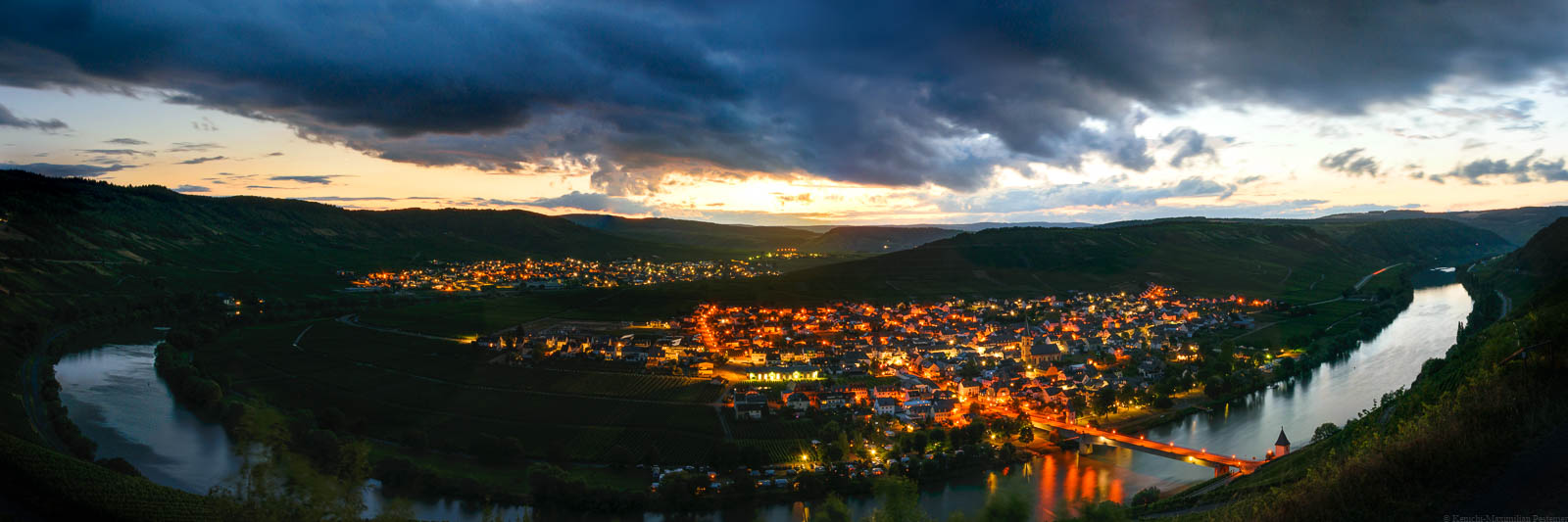 Vom Berg aus blickt man auf die Orte Trittenheim und Leiwen in der Nacht. Die Orte sind aufgrund des Lichts in Orange eingefärbt. Im Fluss Mosel spiegelt sich der stark bewölkte Himmel.