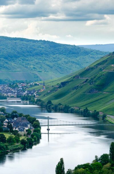 Vom steilen Weinberg Graacher Himmelreich aus blickt man auf den Fluss Mosel im Tal. Am linken Ufer befindet sich der Ort Wehlen. Dieser wird durch eine kleine Stahlbrücke mit dem rechten Ufer verbunden. Dort am rechten Ufer befinden sich die steilen Weinberge Wehlener Sonnenuhr und Zeltinger Sonnenuhr. Im Hintergrund befindet sich der Ort Zeltingen-Rachtig. Am grauen Himmel sind Wolken.