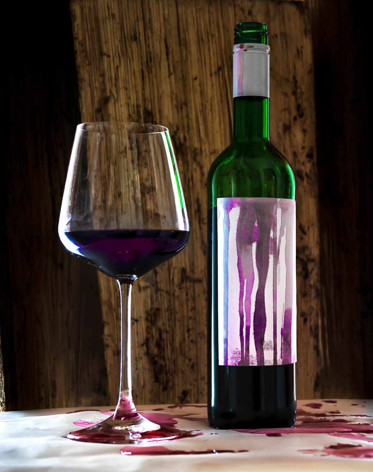 Auf dem Bild ist links ein Glas Rotwein erkennbar. Rechts steht eine Flasche Schomlauer. Das Etikett der Weinflasche und der weiße Papierboden sind von Rotweintropfen bedeckt. Im Hintergrund ist Holz. Das Bild spielt auf Béla Hamvas Werk, "Die Philosophie des Weins" an.