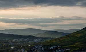 Im Vordergrund befindet sich der Weinbauort Kues. Oberhalb auf der rechten Seite befindet sich der steile Weinberg Kueser Rosenberg. Im Hintergrund fließt die Mosel entlang von weiteren Weinbergen. Am Himmel ist Abendstimmung mit vielen Wolken.