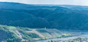 Vorne rechts befindet sich der Ort Piesport. Dahinter fließt der Fluss Mosel. Am anderen Ufer erhebt sich der steile Weinberg Minheimer Günterslay und man erkennt dahinter den Ort Minheim sowie bewaldete Hügel.