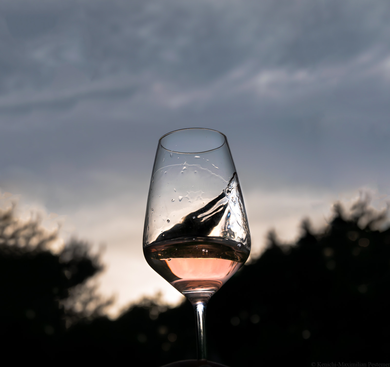 Am Abend wird ein Glas Vinaet Rosé in die Luft gehalten und geschwenkt. Das Glas Rosé befindet sich in der Mitte des Bildes. Im Hinter sind Wälder, Wolken und die Abenddämmerung.