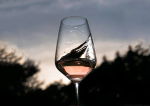 Am Abend wird ein Glas Vinaet Rosé in die Luft gehalten und geschwenkt. Das Glas Rosé befindet sich in der Mitte des Bildes. Im Hinter sind Wälder, Wolken und die Abenddämmerung.
