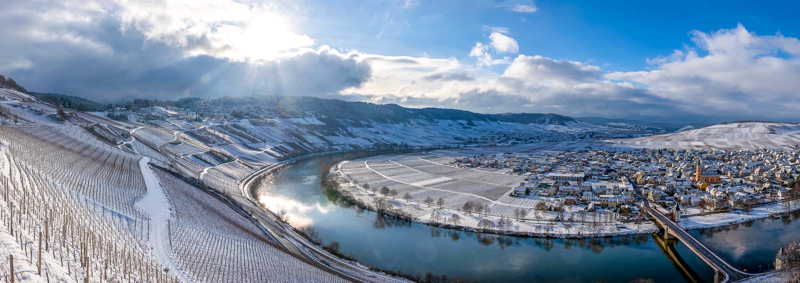 Vom Berg hinab blickt man auf die Moselschleife rund um den Ort Trittenheim im Winter. Der steile Weinberg Trittenheimer Apotheke ist dabei komplett schneebedeckt.
