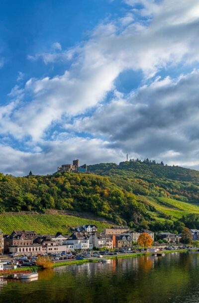 Im Vordergrund fließt der Fluss Mosel. Am linken Flussufer befinden sich Gebäude des Ortes Bernkastel-Kues. Dahinter erhebt sich der Bernkasteler Schlossberg. Es ist ein Weinberg auf dem sich die Burg Landshut befindet. Am blauen Himmel schweben viele Wolken.