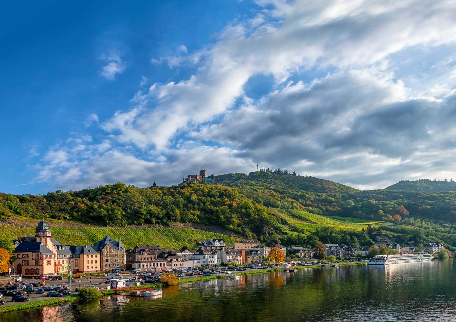 Im Vordergrund fließt der Fluss Mosel. Am linken Flussufer befinden sich Gebäude des Ortes Bernkastel-Kues. Dahinter erhebt sich der Bernkasteler Schlossberg. Es ist ein Weinberg auf dem sich die Burg Landshut befindet. Am blauen Himmel schweben viele Wolken.